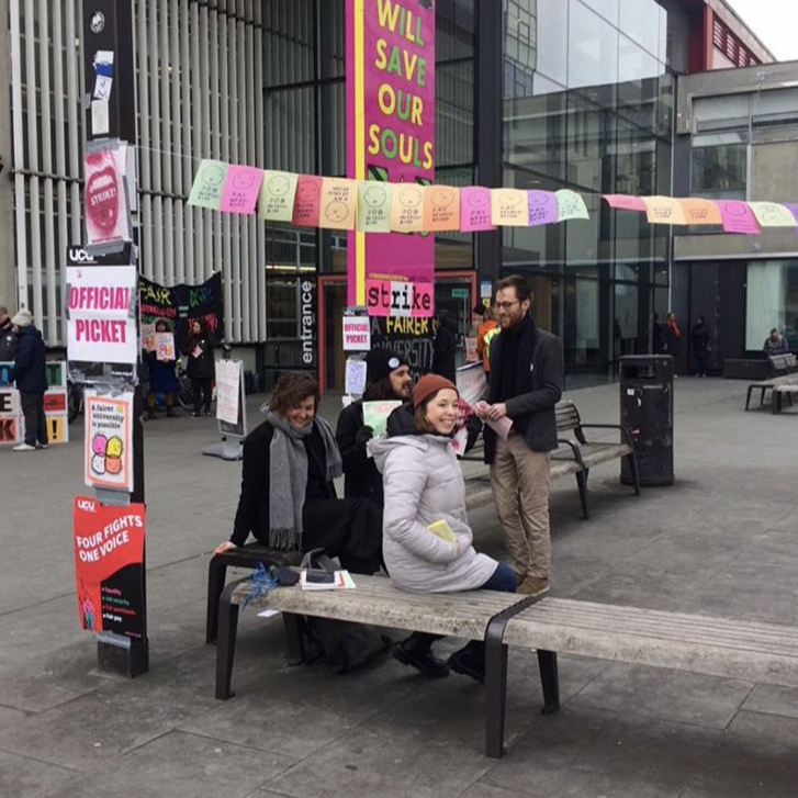 A group of people sitting on benches in front of a university, underneath colourful flags. One sign says "Official Picket" and another says "Strike"