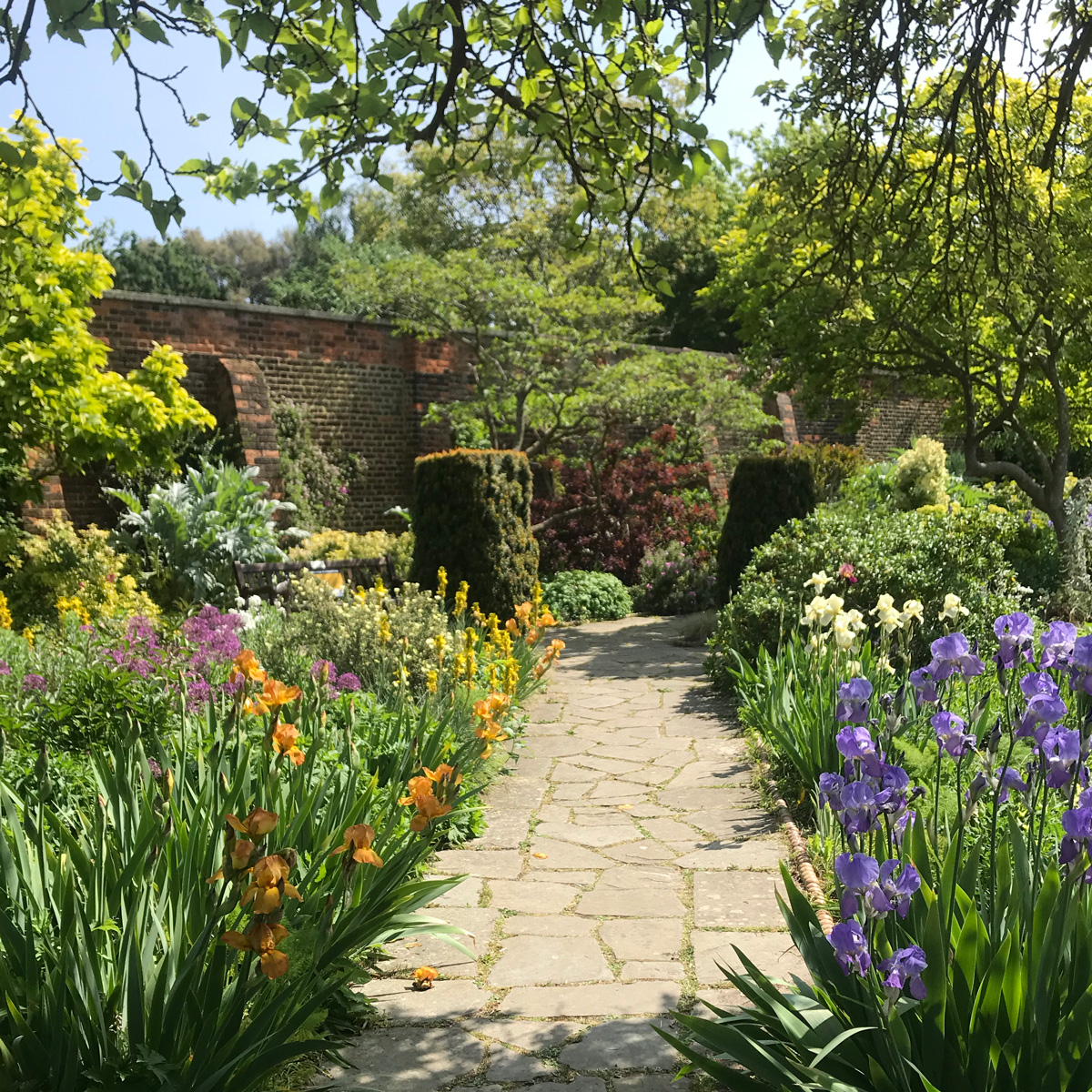 A stone pathway leading into a garden. On either side of the path, are bright flowers in orange, yellow, purple and white.