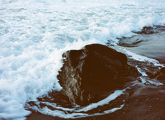 A wave breaking around a rock on a black sand beach