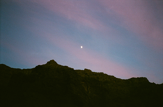 A rocky ridgeline silhouetted against a pink and blue evening sky featuring a gibbous moon
