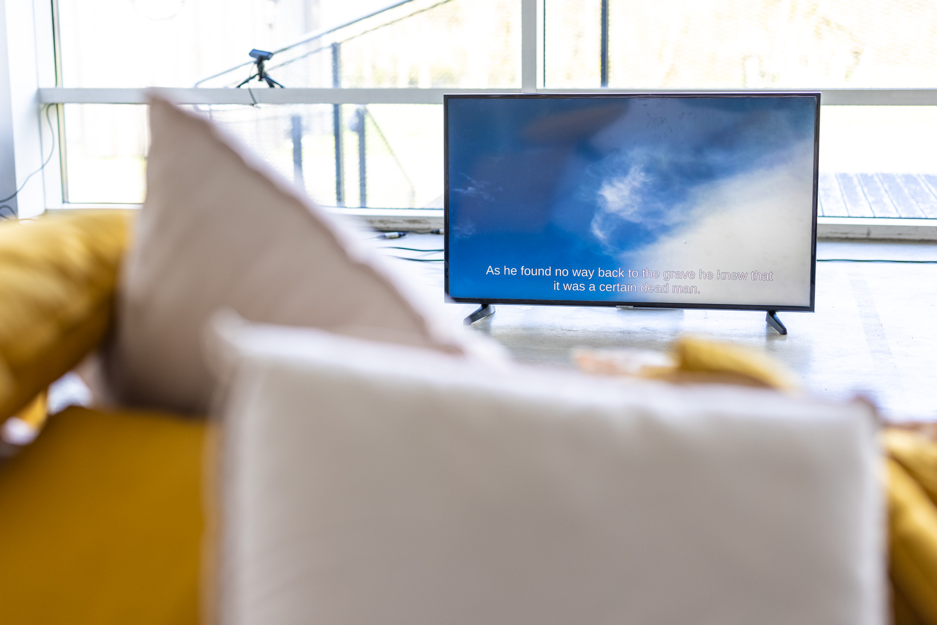 A TV screen in a gallery space showing a live video feed of the sky above. The sky is mainly blue with some wispy clouds. The video is captioned with a sentence that begins with "As he found no way back…".