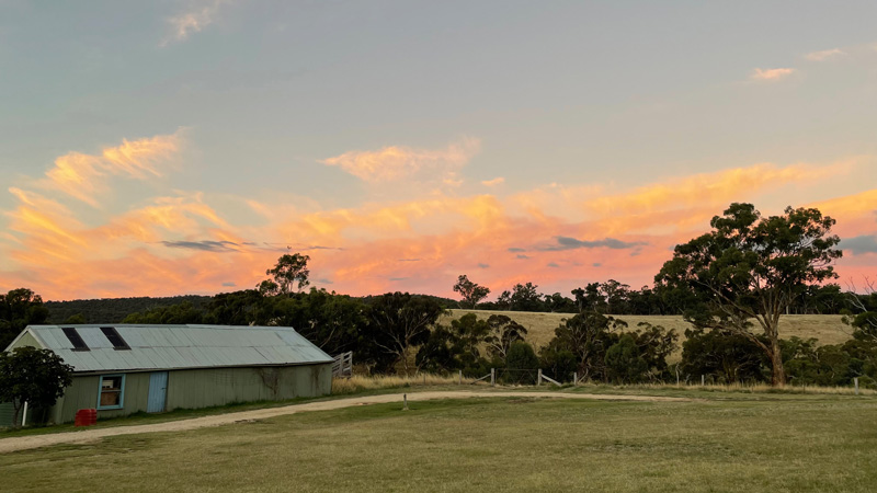A brilliant red and yellow sunset in rural Victoria. There is a farmhouse on the left. The grass looks quite dry and there are some eucalypts along the horizon.