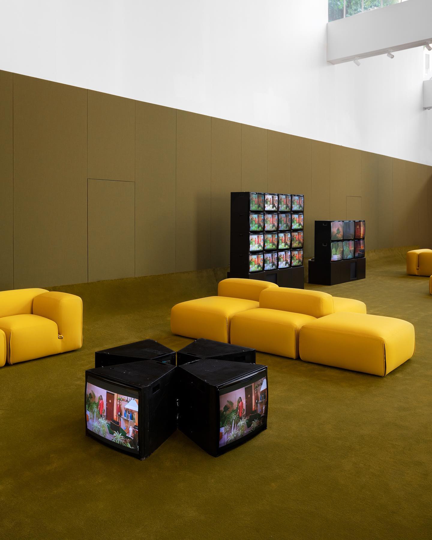 Interior photograph of a contemporary gallery or exhibition space with mustard-yellow modular seating and olive-toned walls.