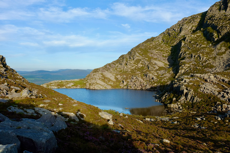 A small lake nestled into the side of a mountain, reflecting the sky. The sky is very blue with some clouds, and there are more mountains in the distance.