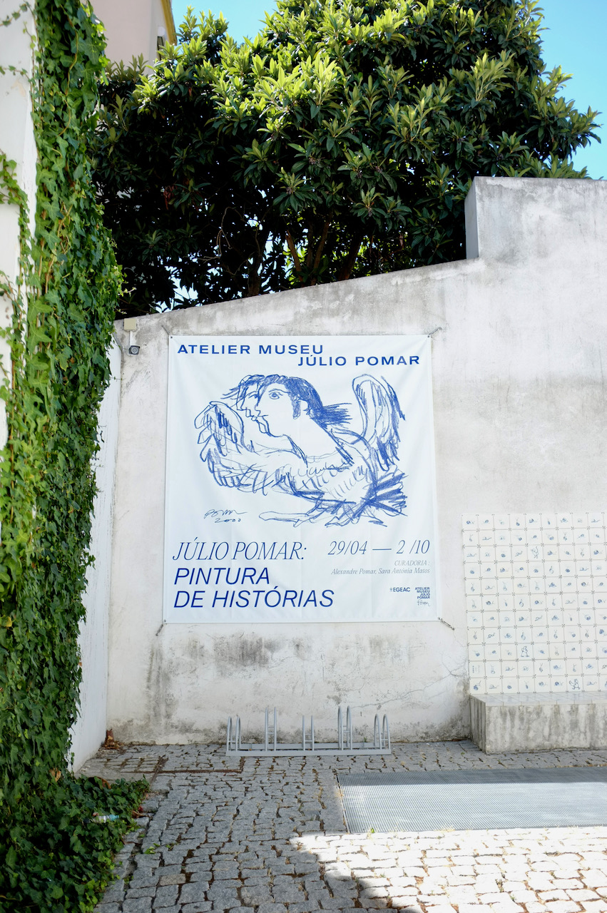 A banner outside the gallery. On one side is a vine-covered wall and on the other are some more tiles.