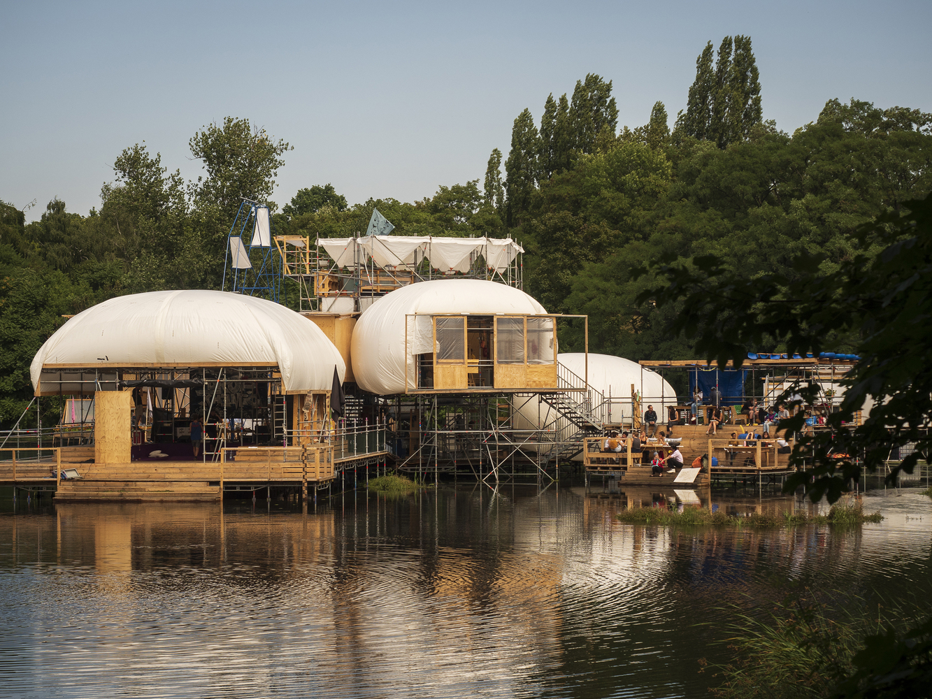 A photo of the Floating University auditorium and kitchen. The structures are made of reclaimed wood and white fabric, perched over the water.