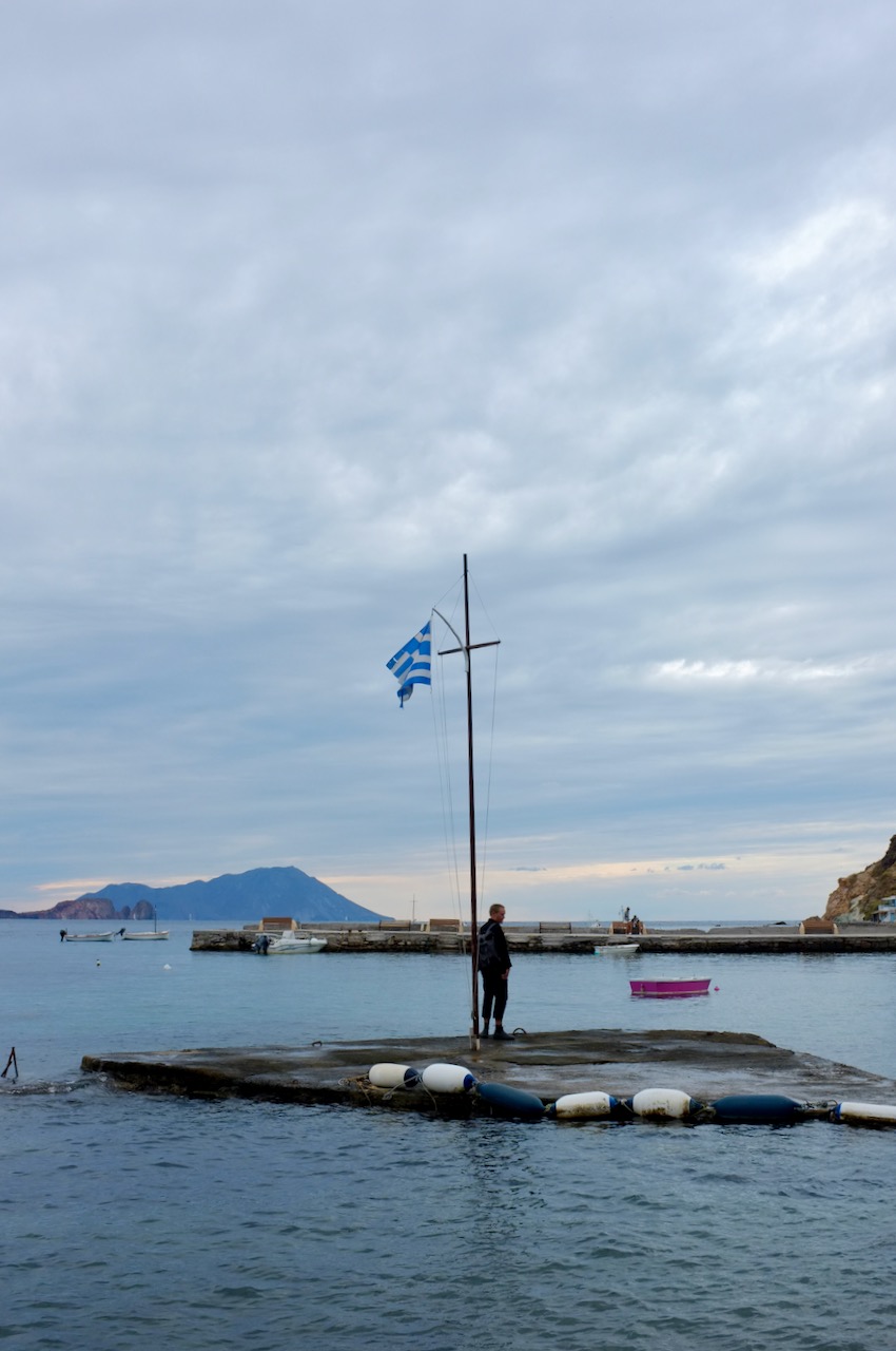 How is standing on a pier, under a Greek flag. The sky is cloudy and everything looks very damp. There are other islands in the background and the sea lapping at my feet.