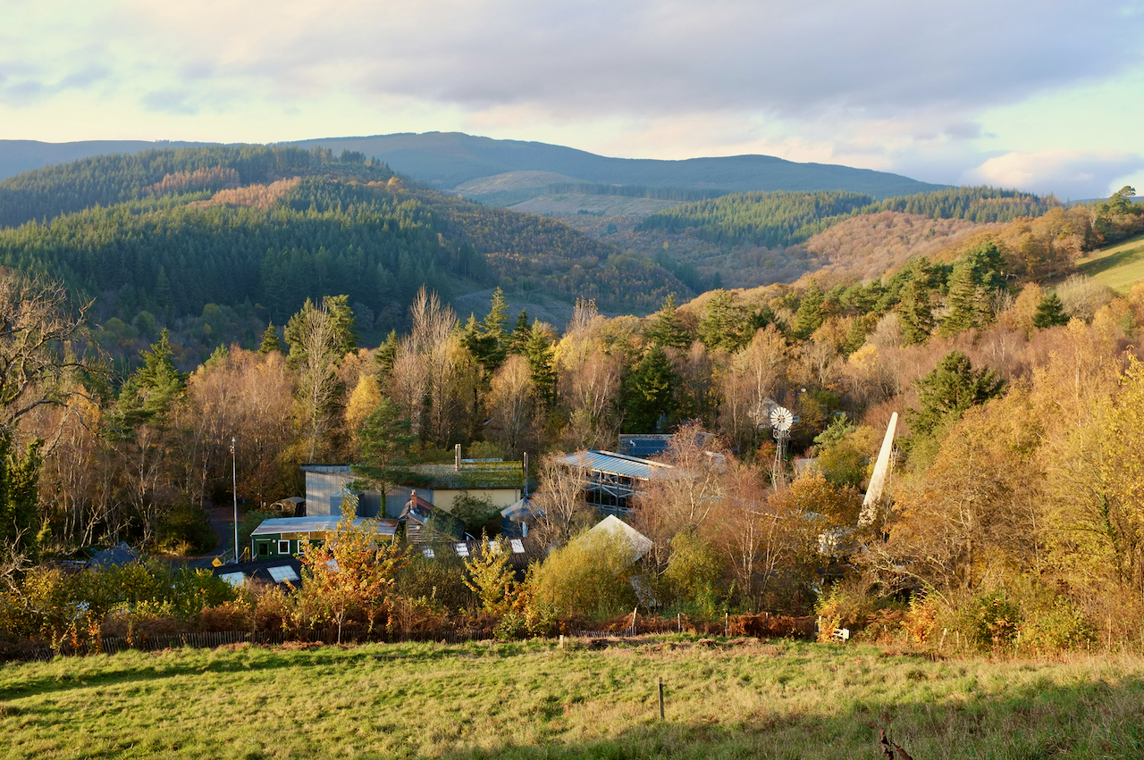 A photo of the Centre for Alternative Technology taken from the hill above. There are a number of usual building structures poking out from amongst the trees, as well as a windmill and a large wind turbine blade on its side. In the background there are hills covered in forest.