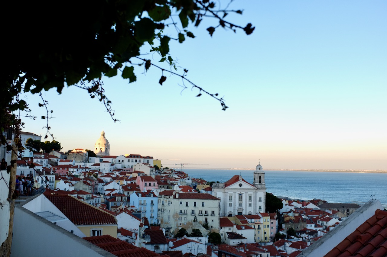 A view of Alfama, with colourful houses dotted on the hillside and the river in the distance. It's sunset and the light is beautifully soft.