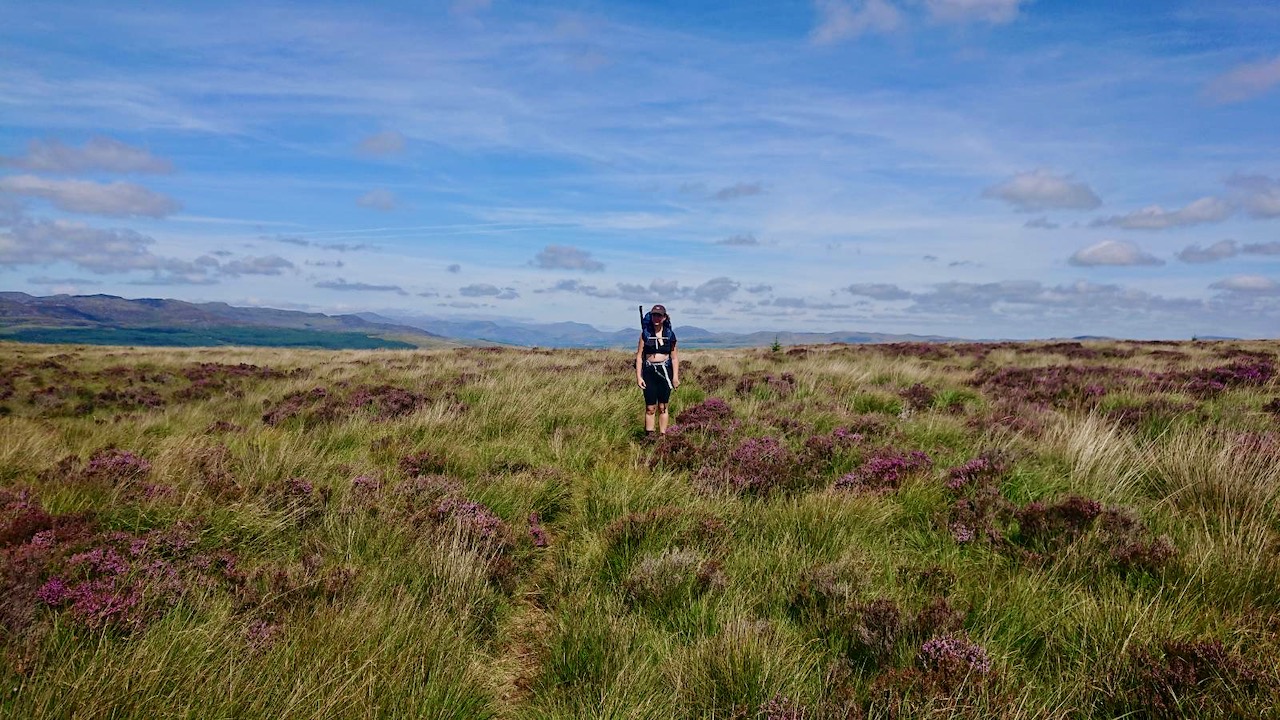 Me standing in a field of heather with a hiking pack on. The sky is blue with some scattered clouds.