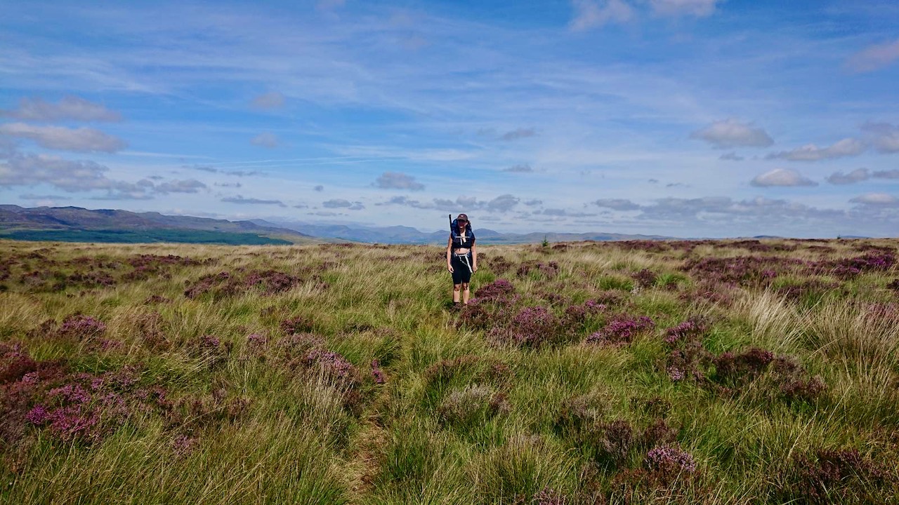 Me standing in a field of heather with a hiking pack on. The sky is blue with some scattered clouds.