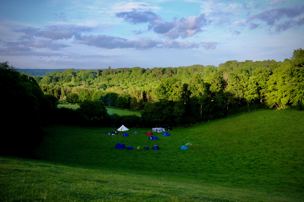 A group of tents at the bottom of a lush green valley at sunset.
