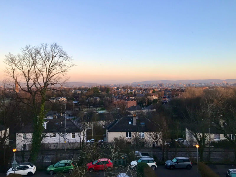 The view from my window on the south side of Glasgow. You can see across the city to the mountains beyond. The sky is a soft gradient from peach to blue.