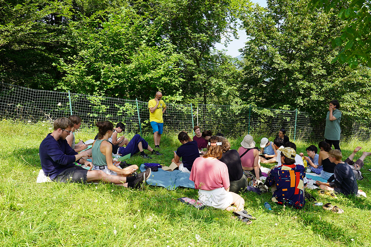 People sitting in the grass, listening to Gianni speak about the commons.