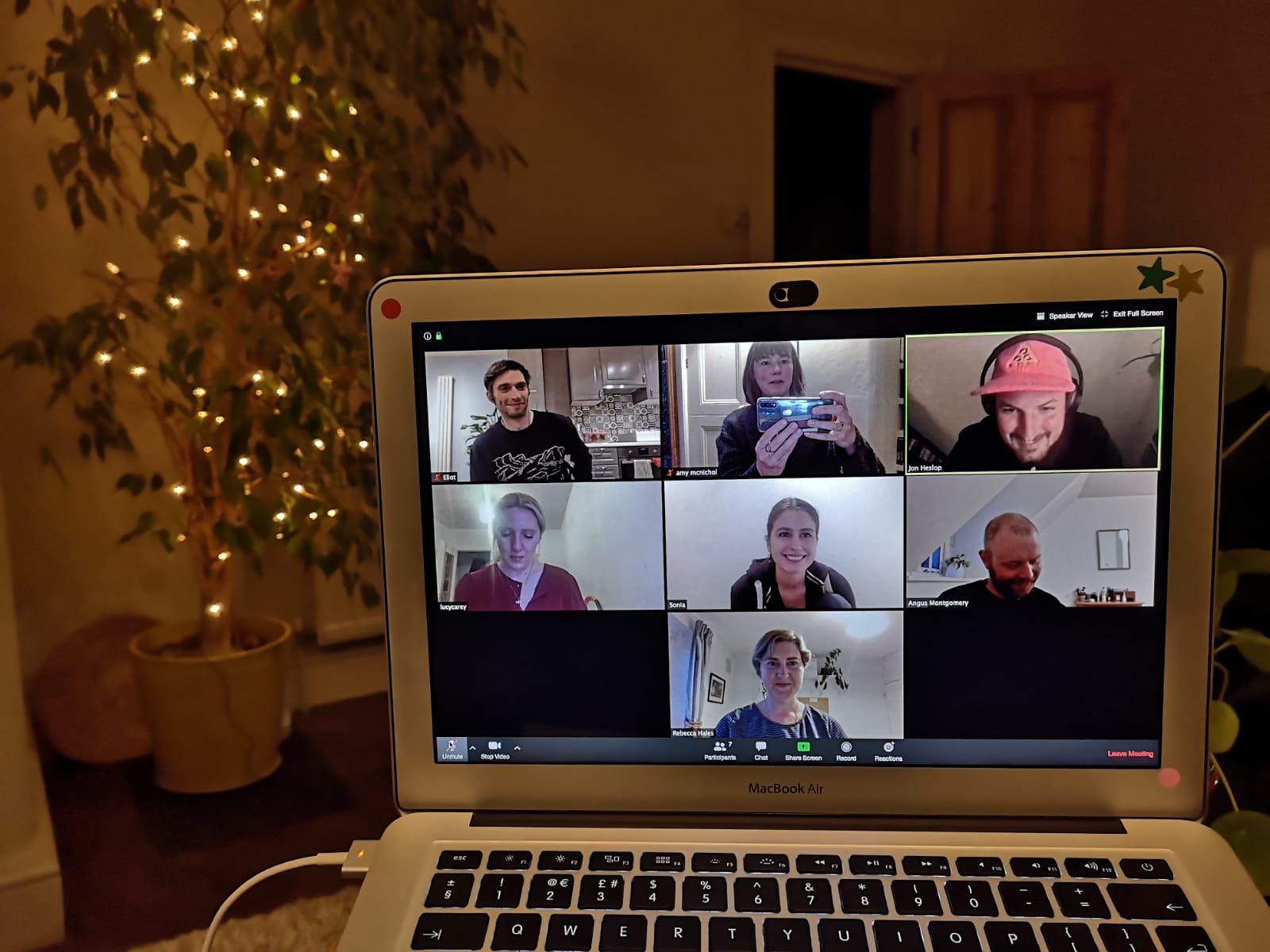 A photo of a computer with a zoom call with Eliot, Amy who is taking the picture, Jon wearing a cap, Lucy, me, Angus, and Becky. Behind the computer is a tree in a pot with fairy lights on it.