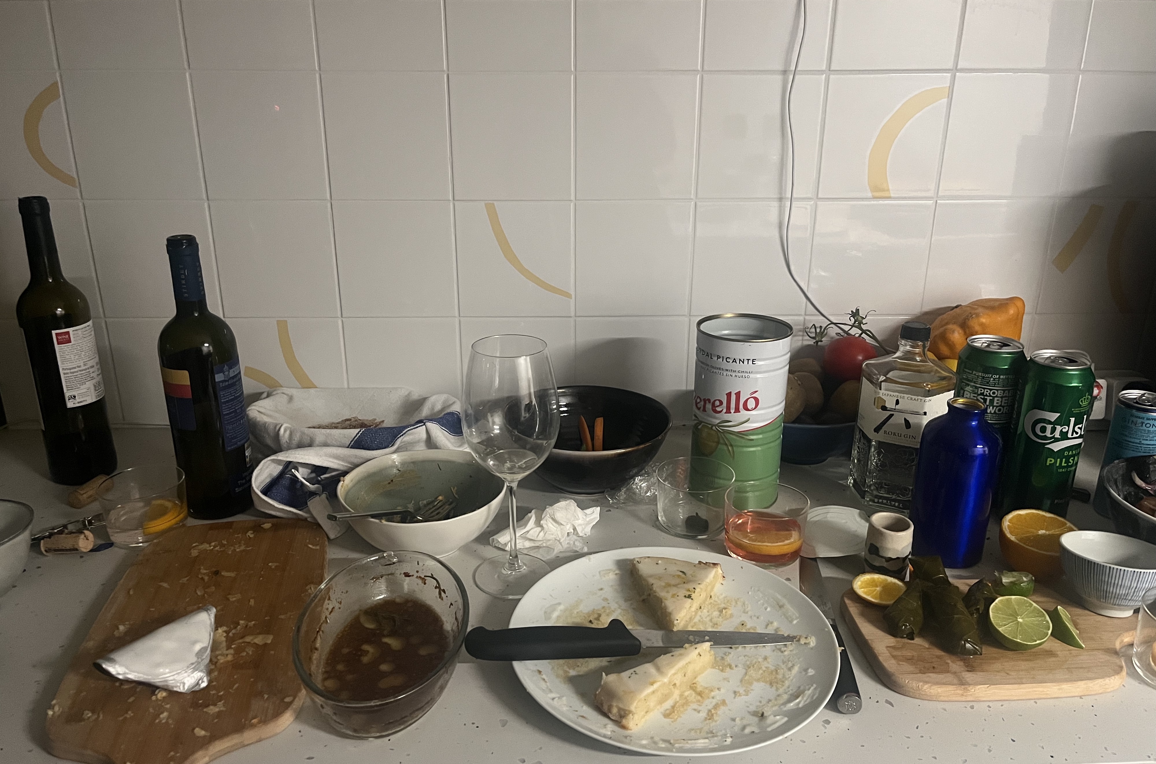 A picture of a kitchen counter covered in party detritus: dirty dishes, 2 empty wine bottles, lager tins, a bottle of Roku gin, a plate with a sliver of cake on it, and a big empty tin of Perello olives