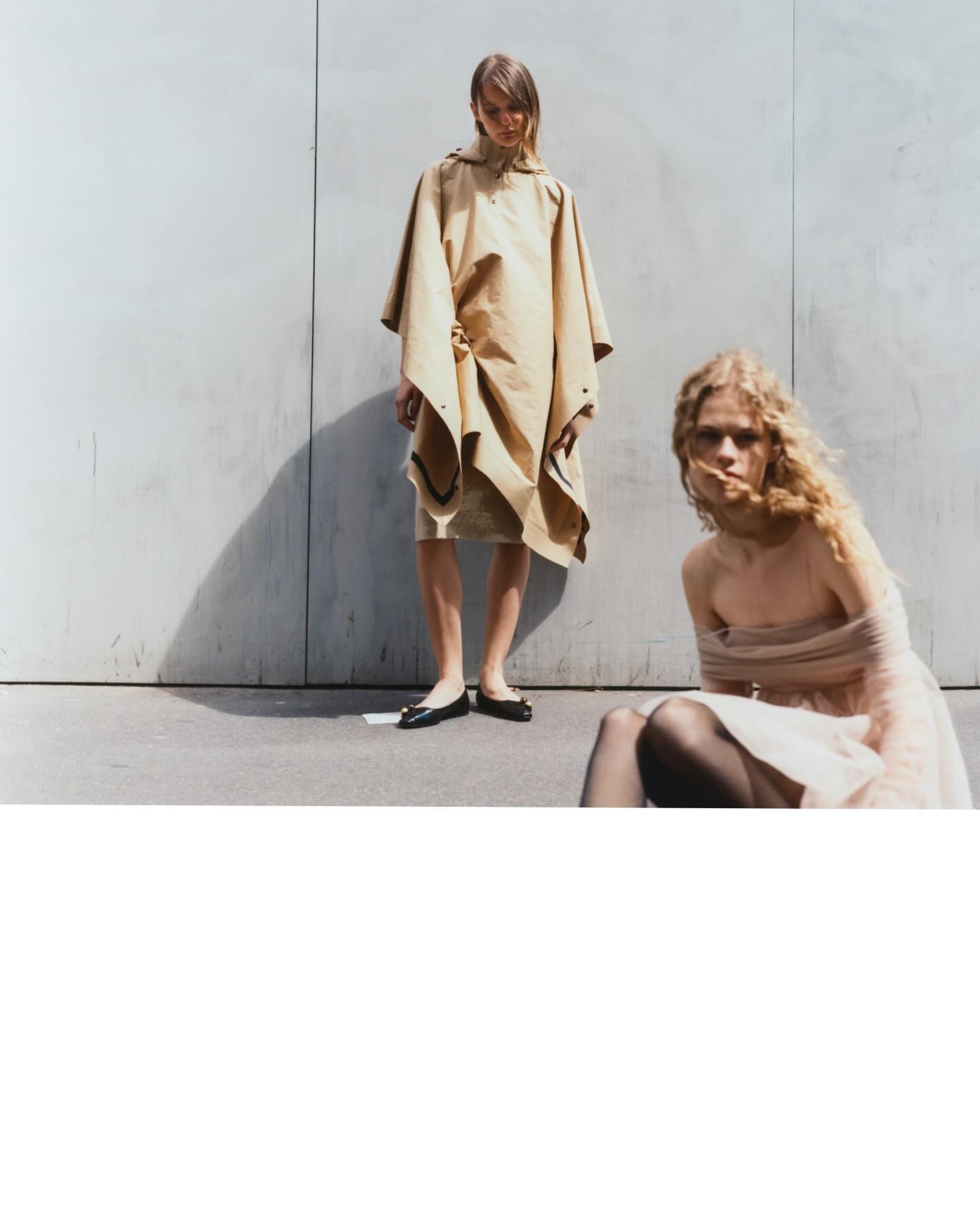 Fashion photography featuring two models against a weathered concrete wall in bright daylight.