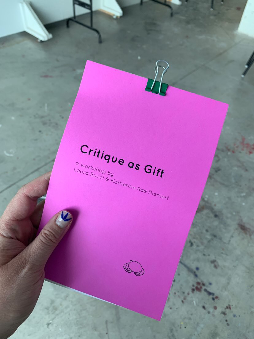 a hand holding a purple booklet with a clip at the top, reads Critique as Gift a workshop by Laura Bucci and Katherine Rae Diemert