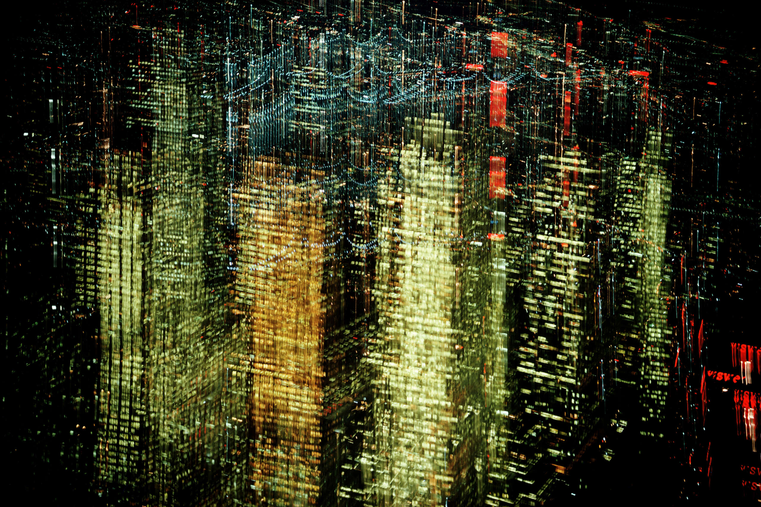 The image displays a vibrant and abstract representation of a cityscape at night. The scene is characterized by streaks of light in yellow, green, and red, giving the impression of motion or reflection. Tall buildings with illuminated windows dominate the view, with the patterns of light and color suggesting distortion or artistic manipulation. The overall effect creates a dynamic, almost dreamlike atmosphere, capturing the energy and complexity of urban life.