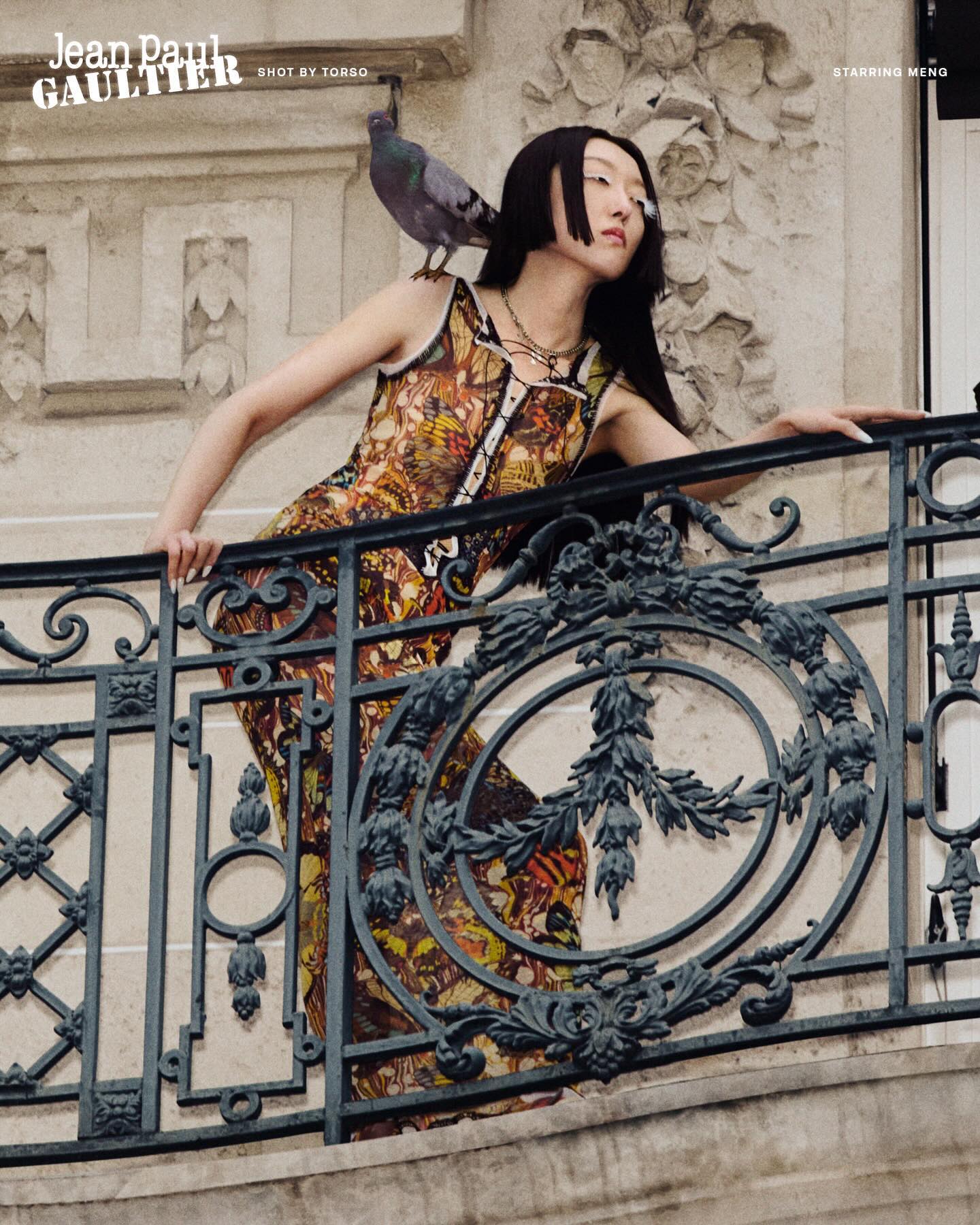 Editorial fashion photograph shot for Jean Paul Gaultier, featuring a model in a gold and rust patterned dress leaning against ornate black wrought-iron balcony railings with a pigeon on her shoulder, set against a cream and beige stone Parisian…