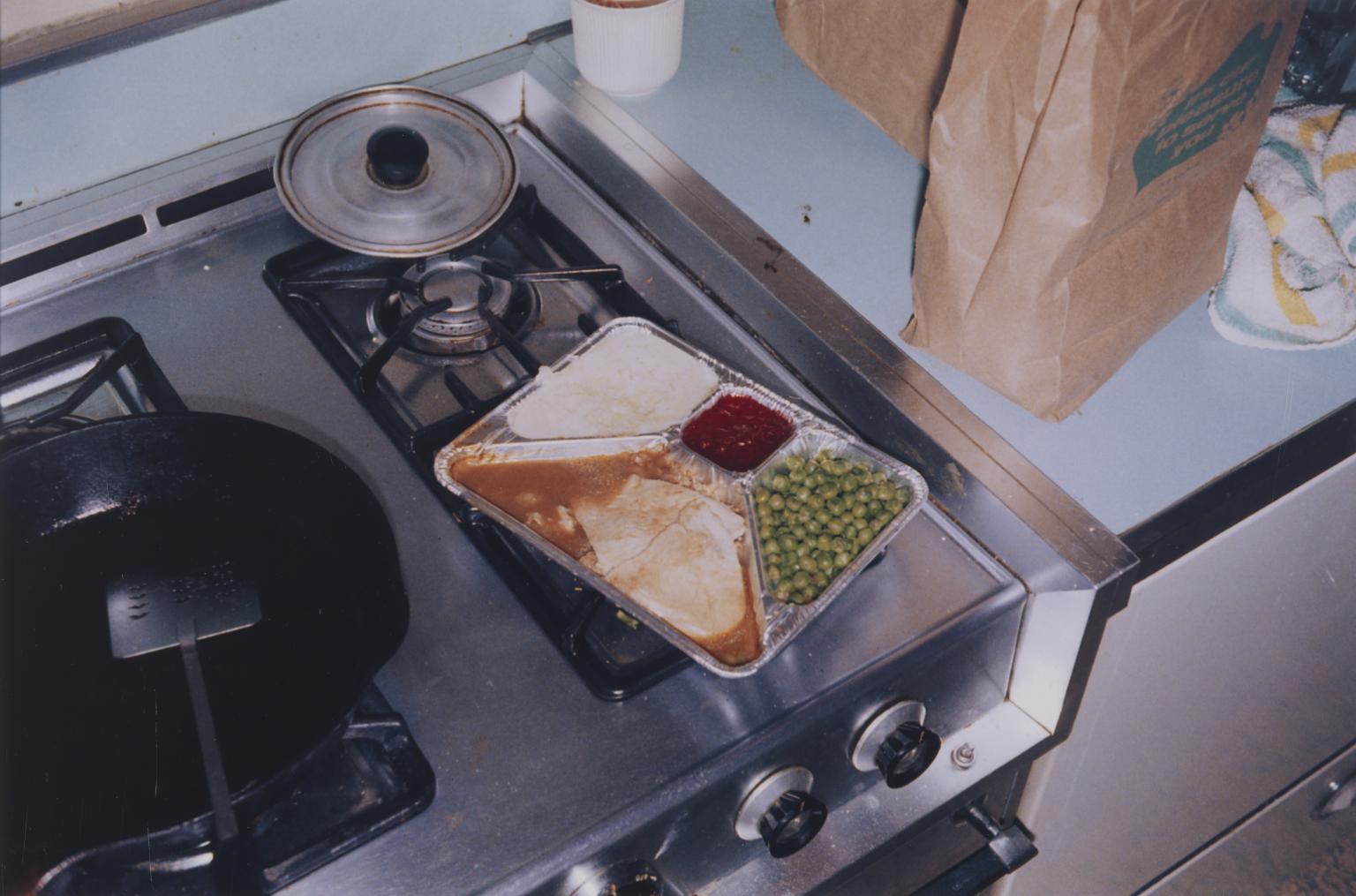 photograph of a TV dinner placed on a stovetop, with a few brown grocery bags on the countertop next to a towel.