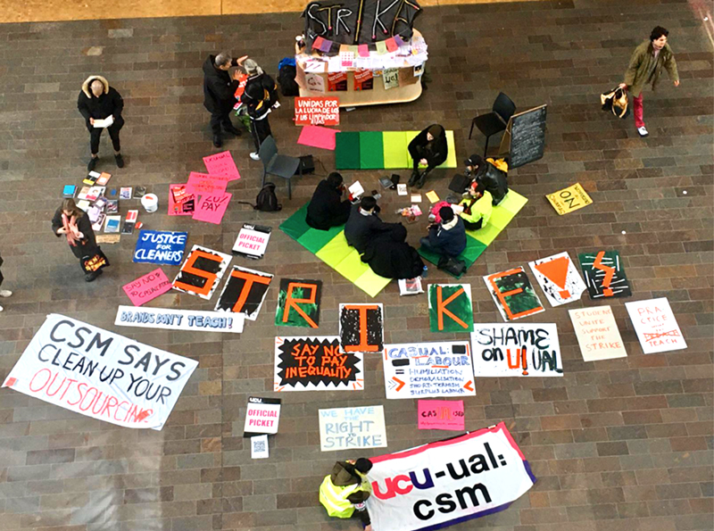 A photo of the CSM picket line, taken from above. There are people sitting on the ground for a teach-out, and colourful signs laid out on the ground.